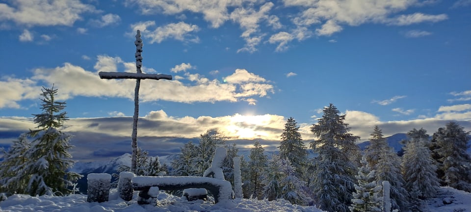 Verschneite Landschaft mit einem Holzkreuz und Bäumen vor Sonnenaufgang.