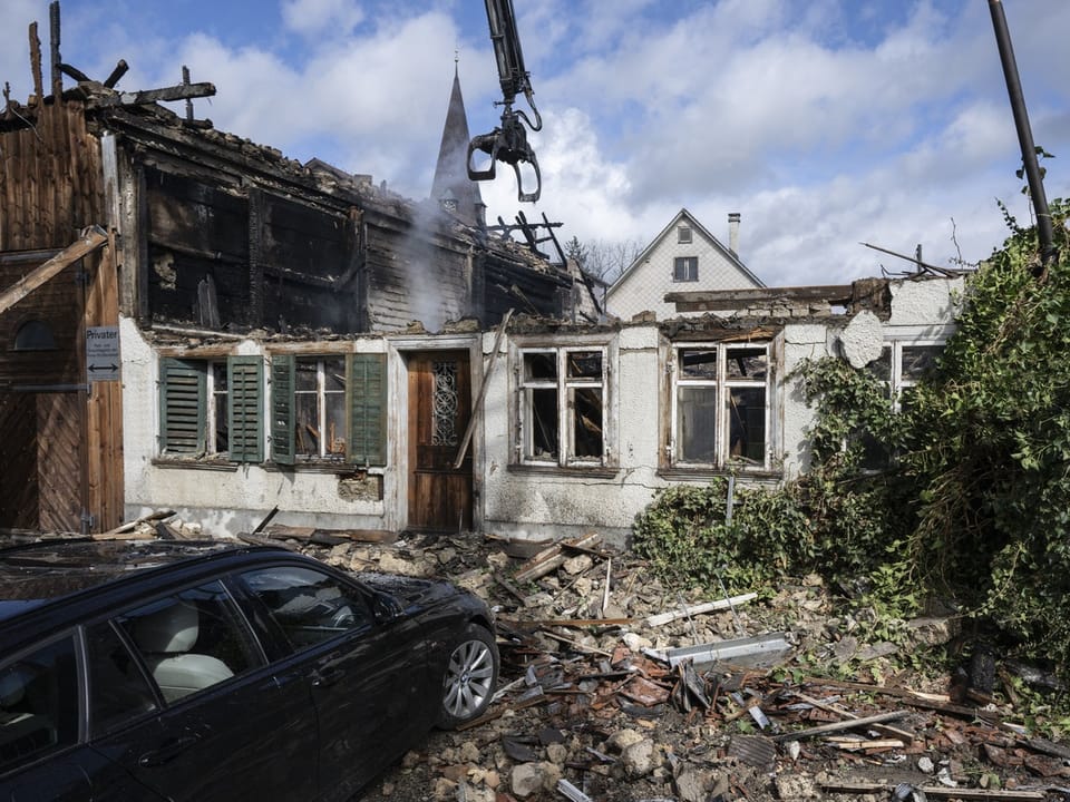 Zerstörtes Haus nach einem Brand mit Trümmern und schwarzem Auto im Vordergrund.