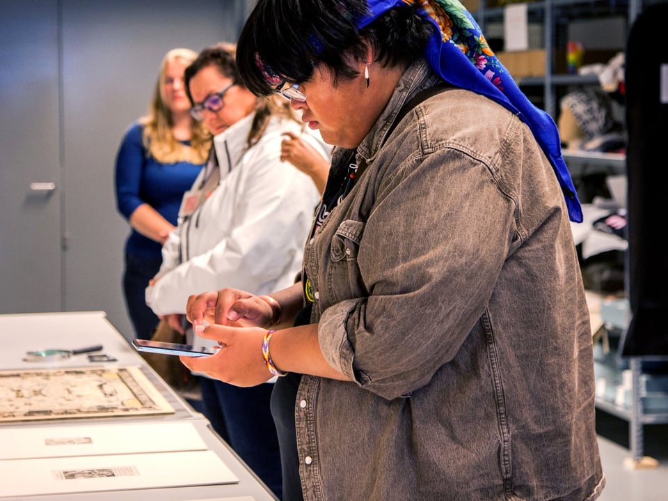 Person betätigt ein Tablet in einer Präsentationsumgebung, andere Personen im Hintergrund.