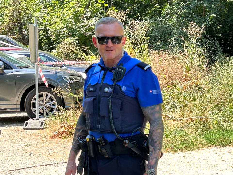 Polizist in Uniform mit Sonnenbrille vor Autos und Büschen.