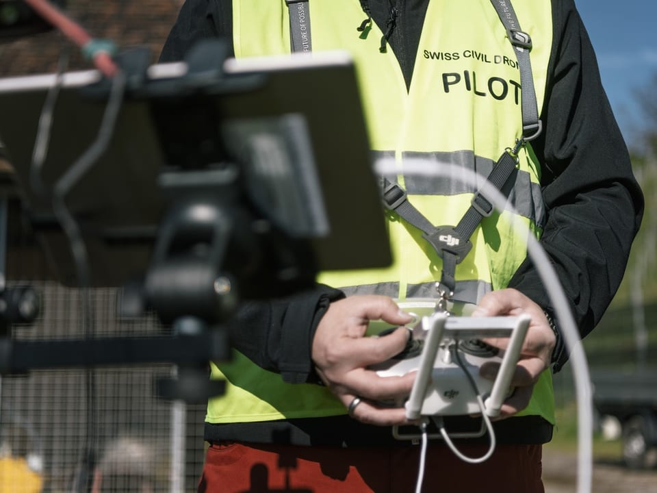 Oberkörper eines Mannes in neongelber Weste mit der Aufschrift «Swiss Civil Drone Pilot» hält Fernbedienung in der Hand.