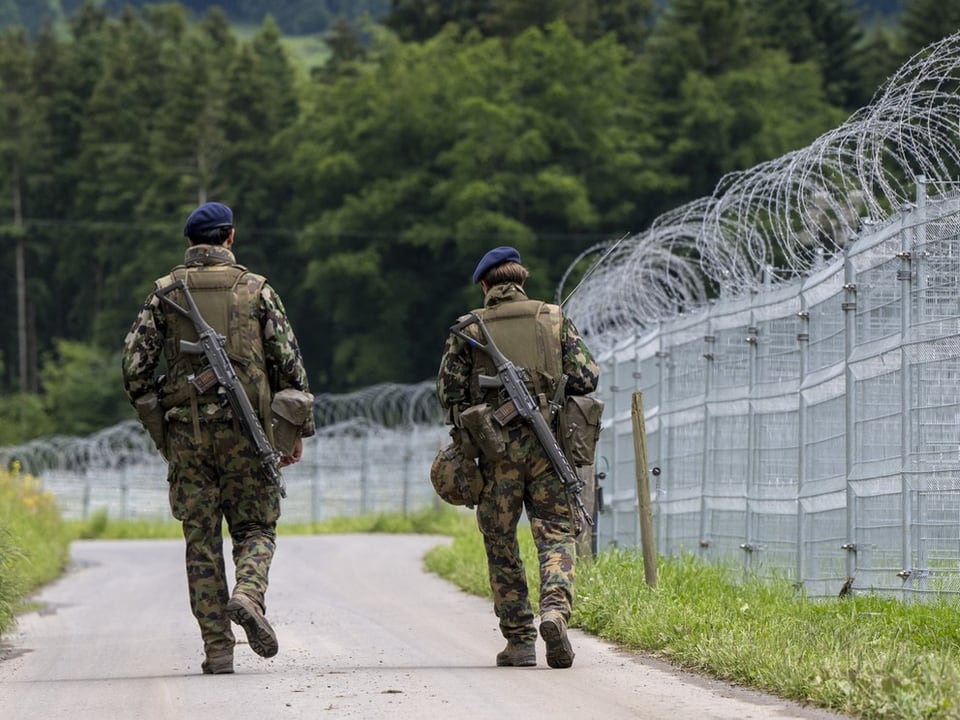 Zwei Soldaten in Uniform patrouillieren neben einem Drahtzaun.