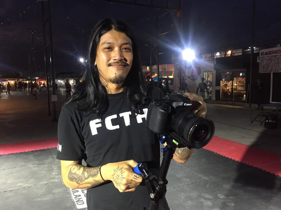 Ein junger Mann mit langen Haaren und einer Profi-Kamera in der Hand. Er trägt ein schwarzes T-Shirt mit dem Logo FCTH (Fight Club Thailand).