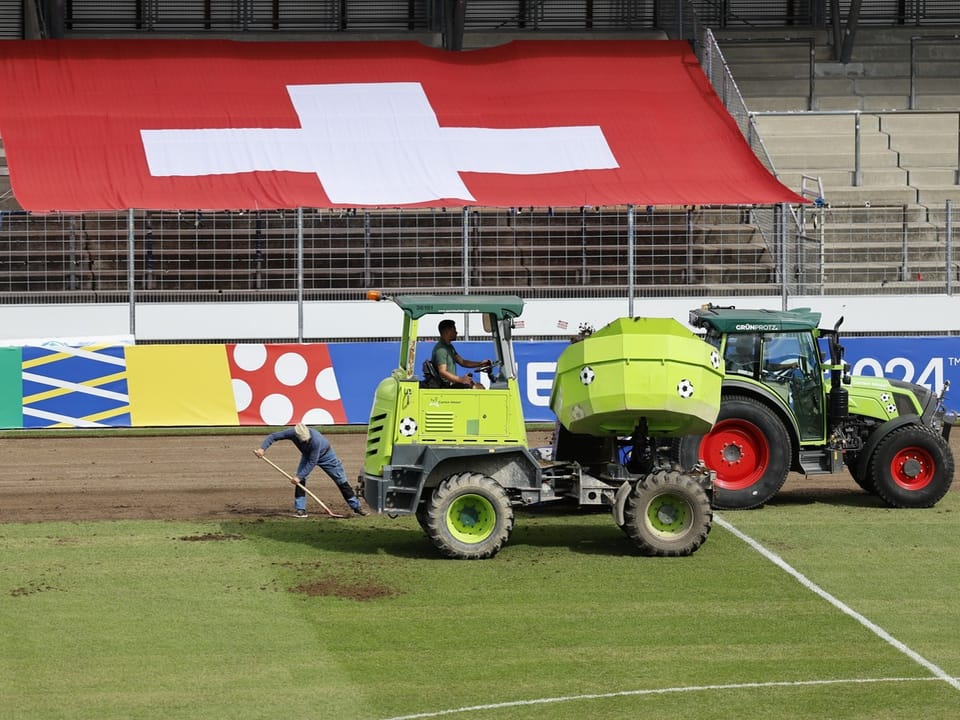 Traktoren und Arbeiter auf einem Fussballfeld vor einer Schweizer Flagge.