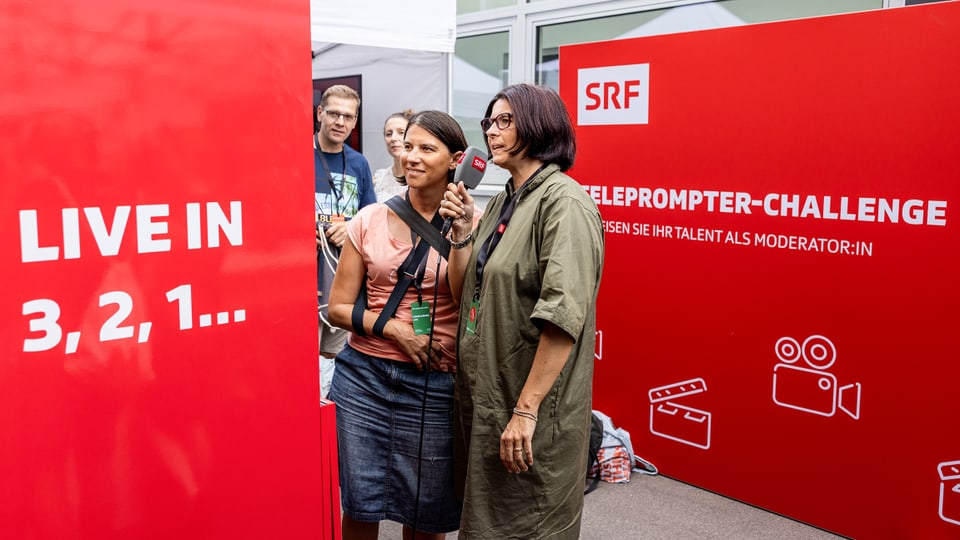 Zwei Frauen halten ein Mikro in der Hand und versuchen, Text vom Teleprompter abzulesen.