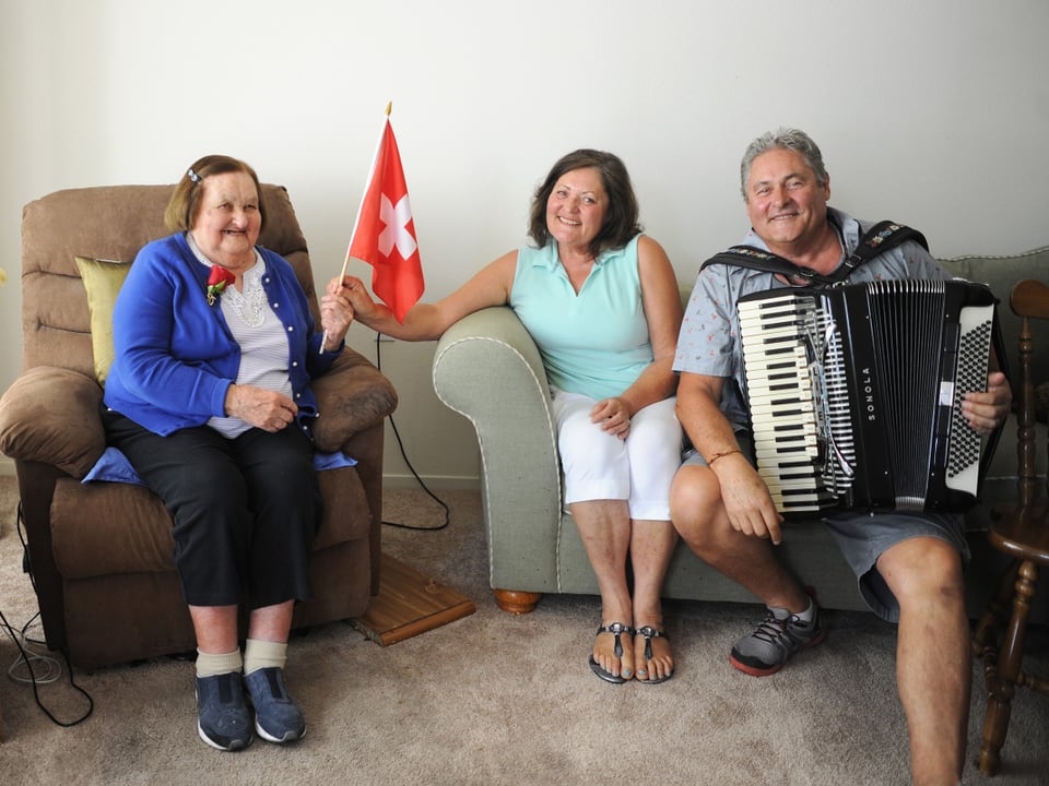 Drei lachende Menschen, eine Frau hält eine Schweizer Flagge, ein Mann spielt Akkordeon.