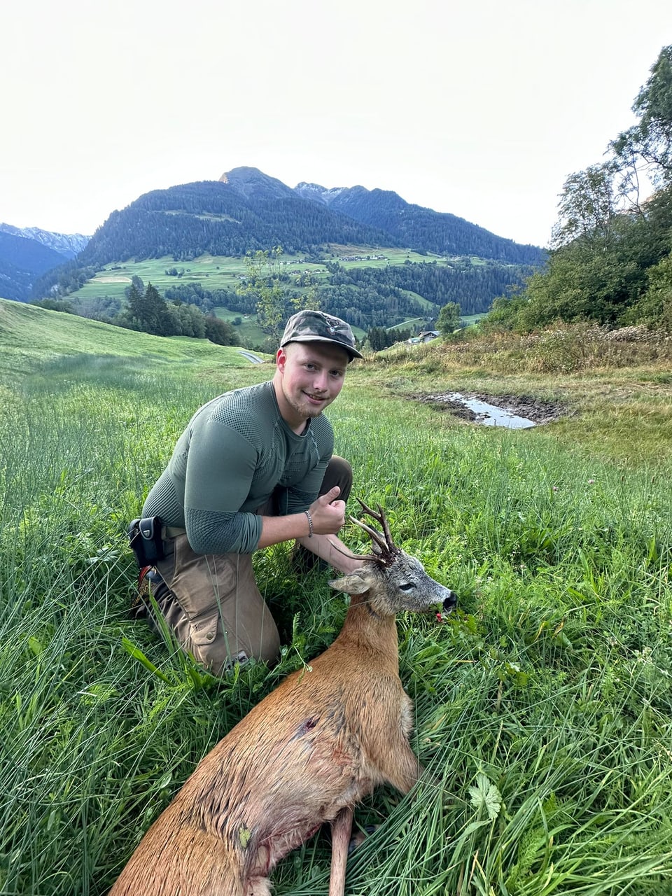 Jäger mit erlegtem Reh im Grünen vor Berglandschaft.