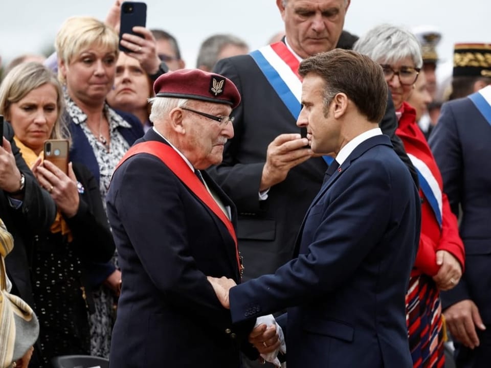 Achille Muller mit einem Beret schüttelt Emmanuel Macron die Hand.
