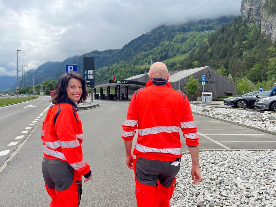 Mona und ein Mann laufen auf die Gotthard Raststätte zu. Sie tragen leucht-orange Arbeitskleidung.