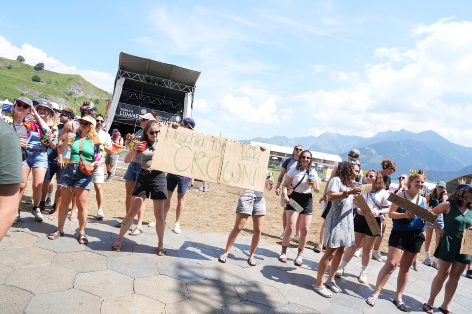 Menschenmenge bei einem Outdoor-Konzert mit Plakat und Bergen im Hintergrund.