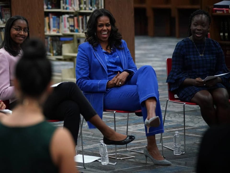Michelle Obama sitzt in einer Bibliotheck auf einem Stuhl. Rundherum sitzen andere Frauen auf Stühlen.
