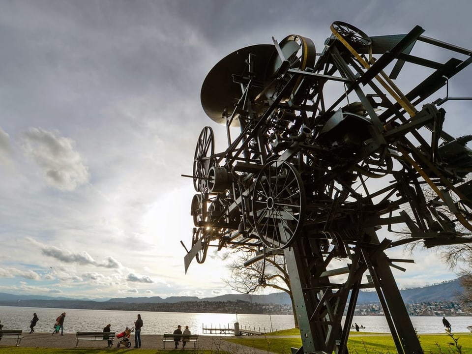 Mechanische Skulptur am Seeufer mit Spaziergängern und Wolkenhimmel.