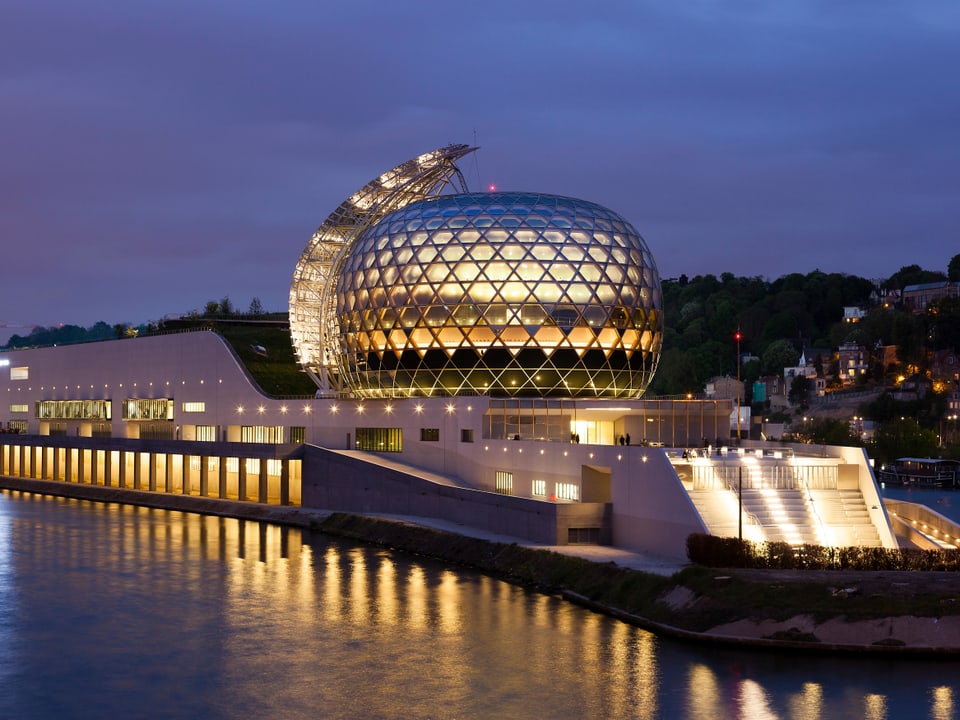 Rundes Glasgebäude steht auf einer Insel in einem Fluss und ist von innen erleuchtet.