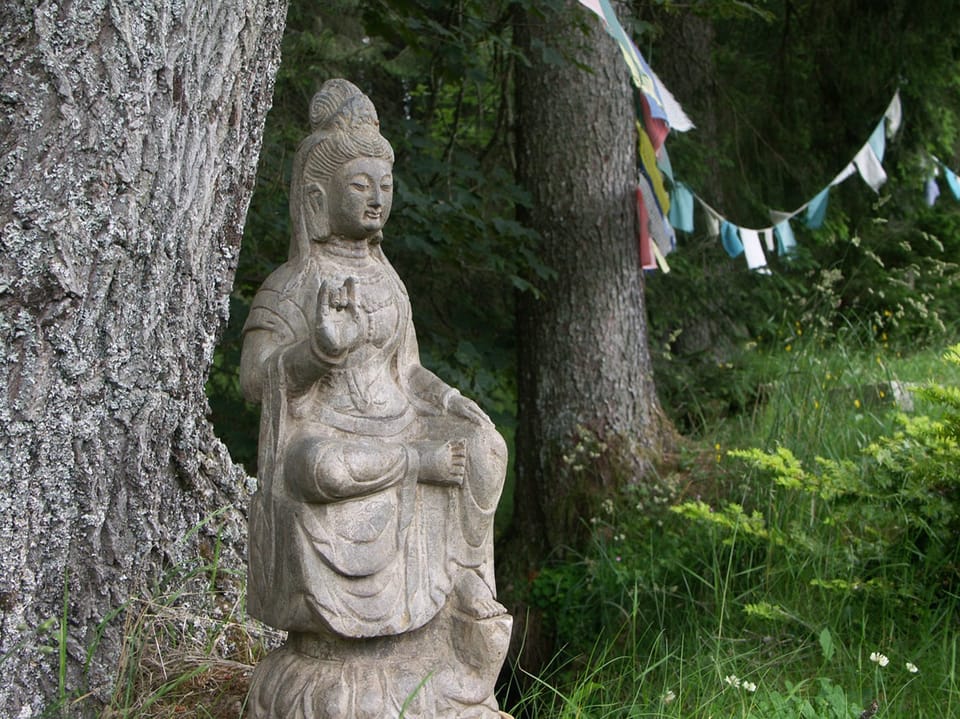 Steinstatue einer sitzenden Buddha-Figur im Wald, neben Bäumen und Gebetsfahnen.