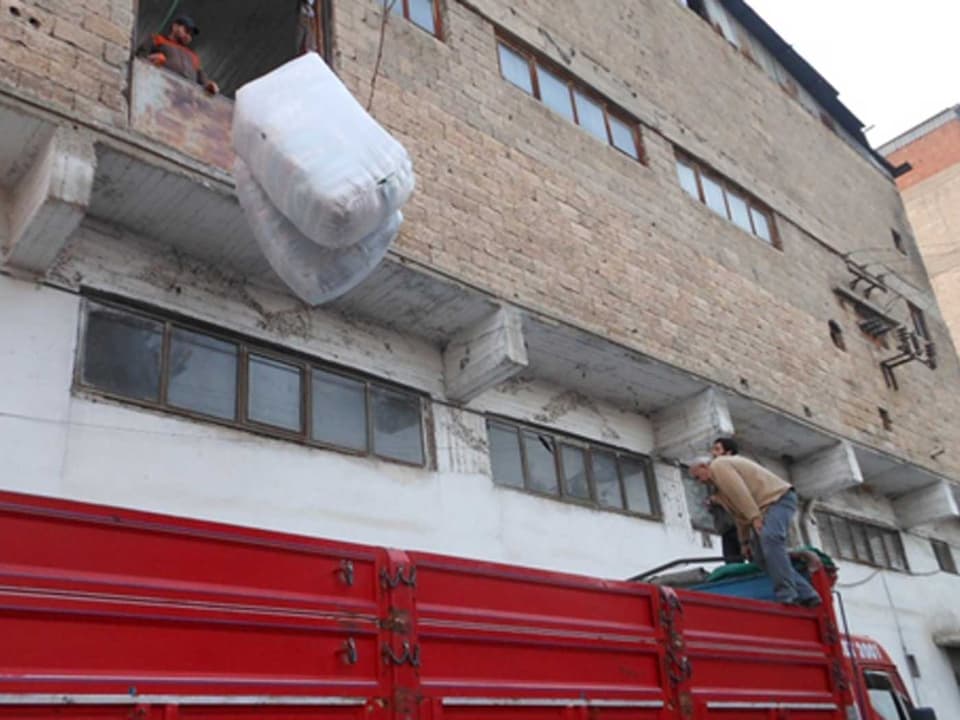 Zu sehen ist ein Mann, der zwei grosse Packen mit Decken aus einem Fenster im ersten Stock wirft. Die Packen werden in einem roten Lastwagen landen.
