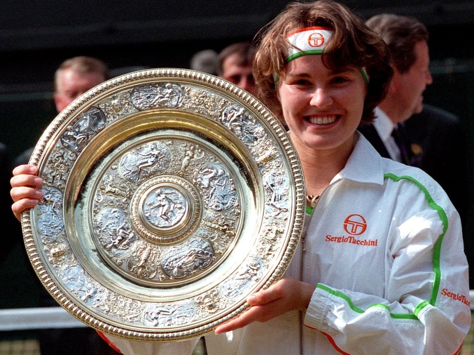 Martina Hingis hält Tennistrophäe im weissen Outfit.