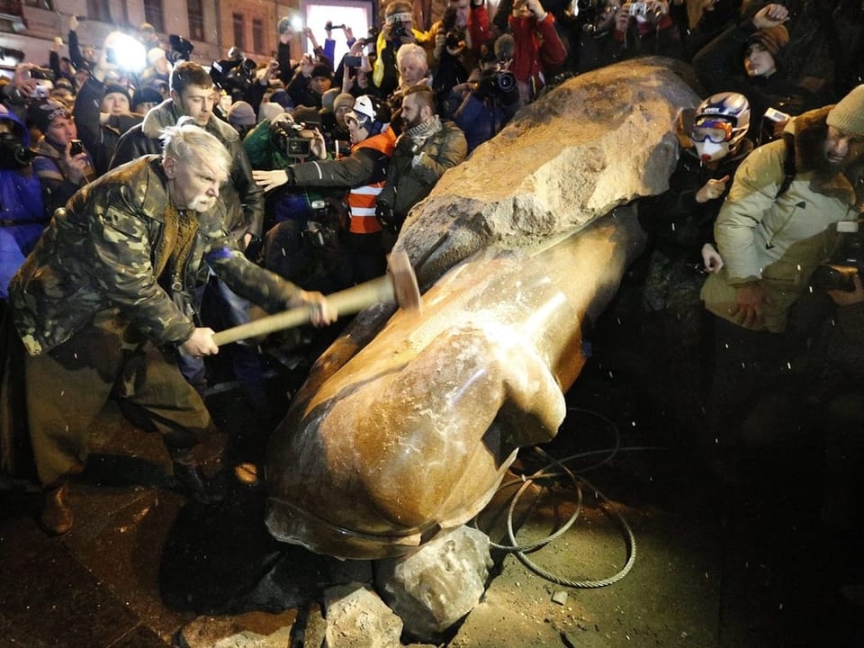 Foto einer Menschenmenge, ein Mann schälgt gerade mit Hammer auf steinerne, gestürzte Statue ohne Kopf