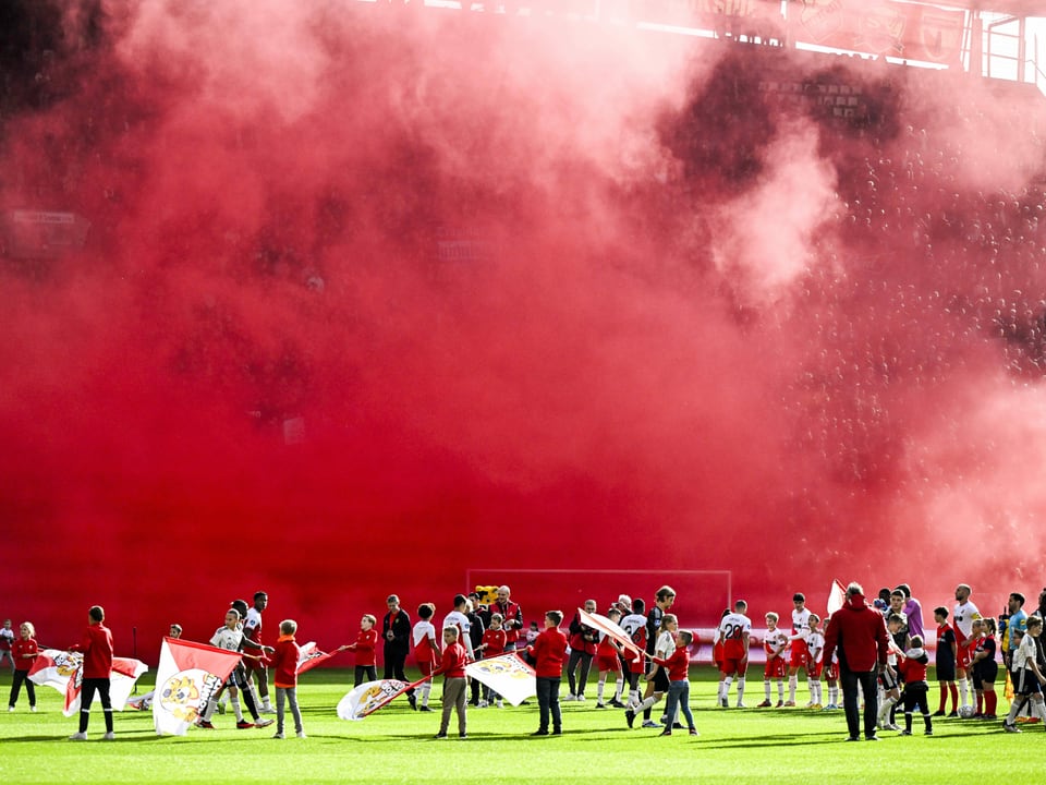 Rote Rauchwolken im Stadion im Hintergrund, vorne schwenken Kinder Fahnen