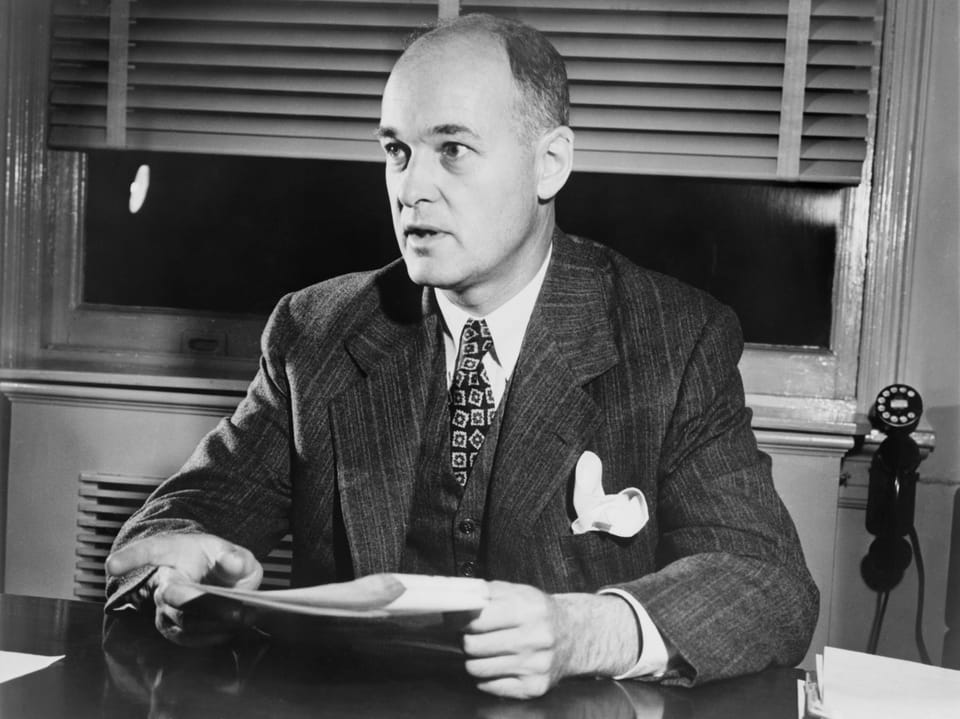 Schwarzweissfoto eines Mannes im Anzug, der an einem Schreibtisch sitzt.