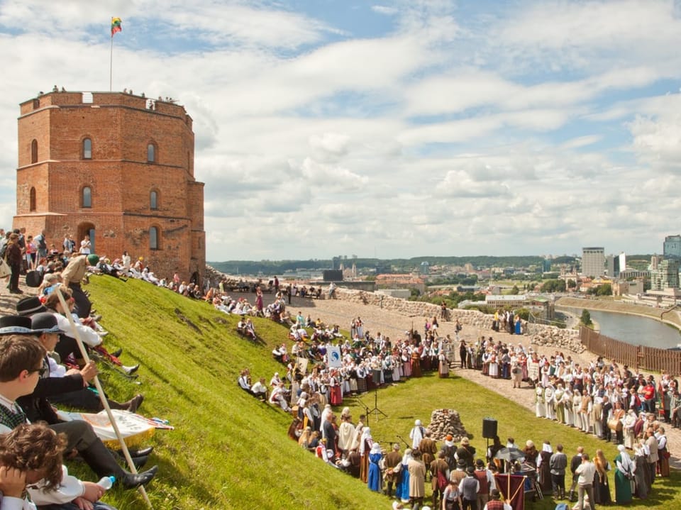 Menschen in traditioneller Kleidung versammeln sich auf einem Hügel vor einem Turm.