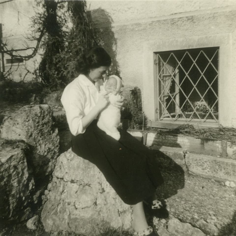 Frau sitzt auf einem Stein und hält liebevoll ein Baby, neben einem Haus mit Gitterfenster.