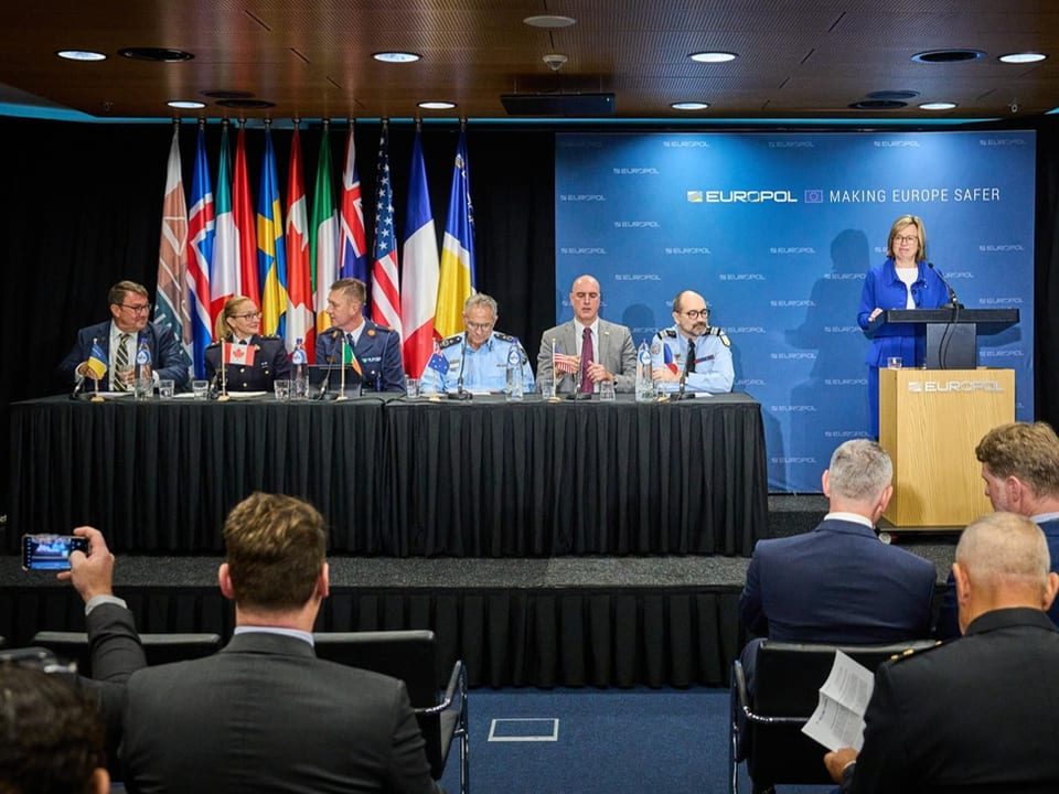 Europol-Pressekonferenz mit sechs Personen an einem Tisch und einer Rednerin am Podium, Nationalflaggen im Hintergrund.