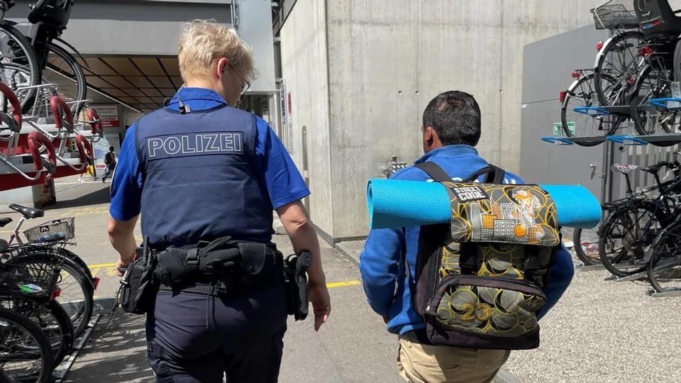 Eine Polizistin und ein Mann mit Rucksack und Iso-Matte laufen Richtung Bahnhof. 