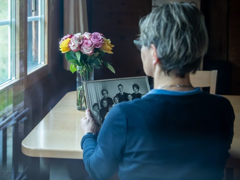 Rückansicht: Eine Frau sitzt an einem Tisch mit Rosen und betrachtet eine Schwarzweiss-Aufnahme