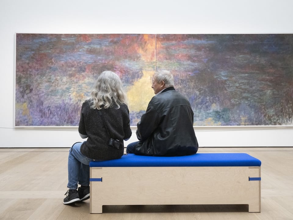 Zwei Personen sitzen auf einer Bank vor einem Gemälde und sprechen miteinander.