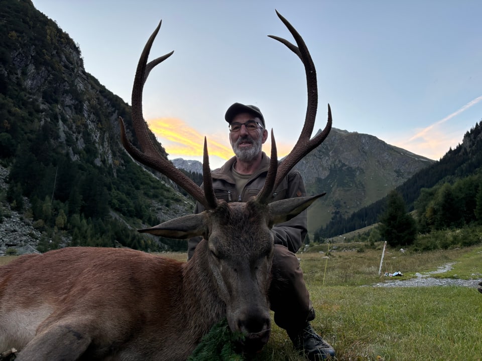 Mann posiert mit erlegtem Hirsch in einer Berglandschaft.