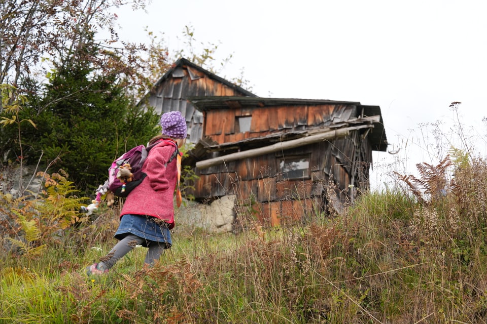 Kind mit Mütze geht einen Hügel hinauf zu einer alten Holzhütte.