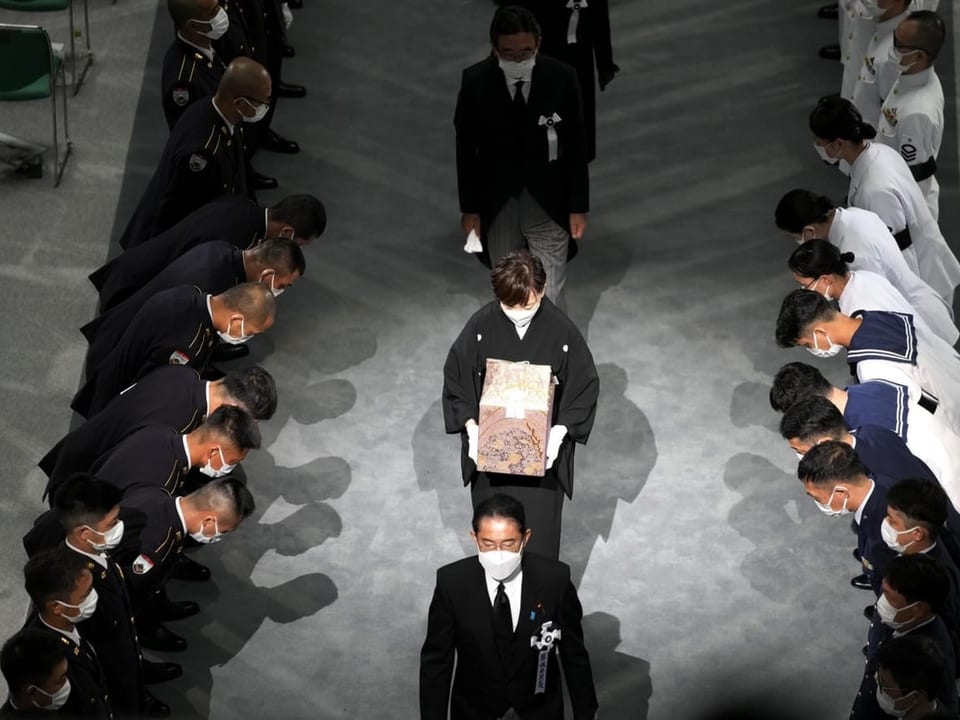 Shinzo Abes Frau trägt seine Urne und geht flankiert von sich verbeugenden Menschen einen Gang entlang.