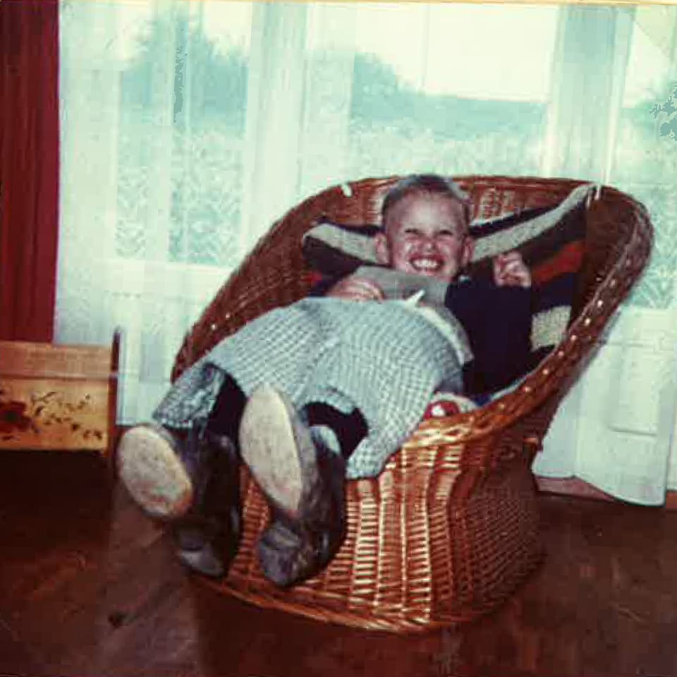 Ein Bub sitzt mit einem Rock und Damenschuhen in einem Korbsessel.