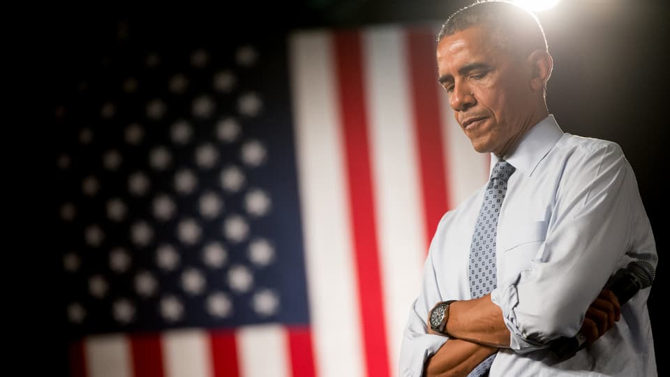 Obama steht mit verschränkten Armen und nachdenklichem Blick nach unten seitwärts vor einer US-Flagge, die im Hintergrund aufgehähngt ist.