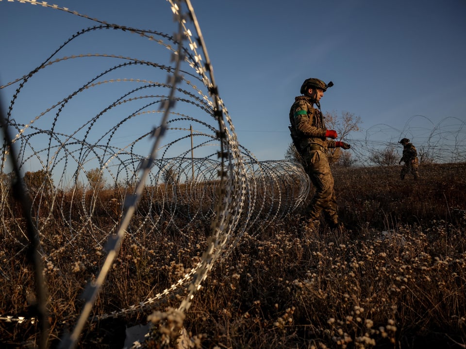 Soldaten patrouillieren entlang Stacheldrahtzaun in Landschaft.