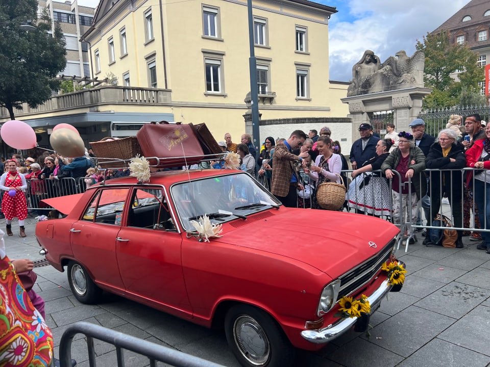 Rotes Auto mit Koffer auf dem Dach, umringt von Zuschauern bei einer Veranstaltung.