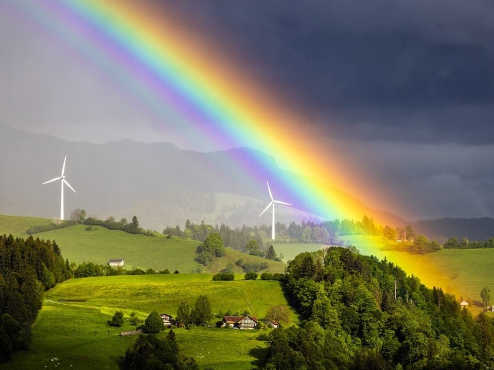 Regenbogen über hügeliger Landschaft mit Windrädern.