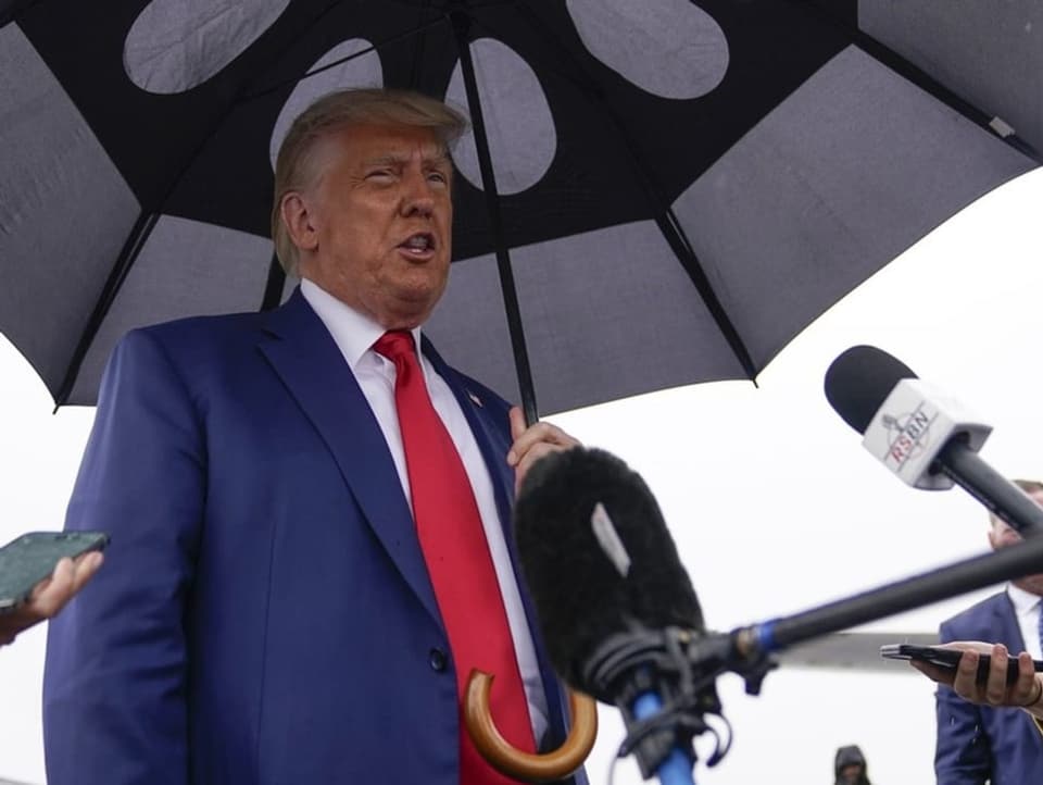 Donald Trump unter einem Regenschirm. Viele Mikrophone.
