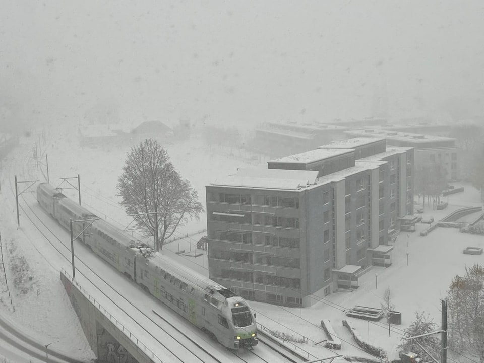 Zug fährt bei starkem Schneefall an Wohnblock vorbei.