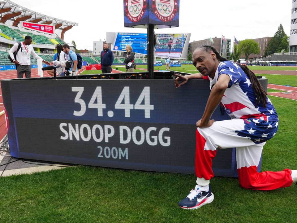 Mann posiert neben einer Anzeigetafel mit der Aufschrift '34.44 SNOOP DOGG 200M' auf einem Sportfeld.