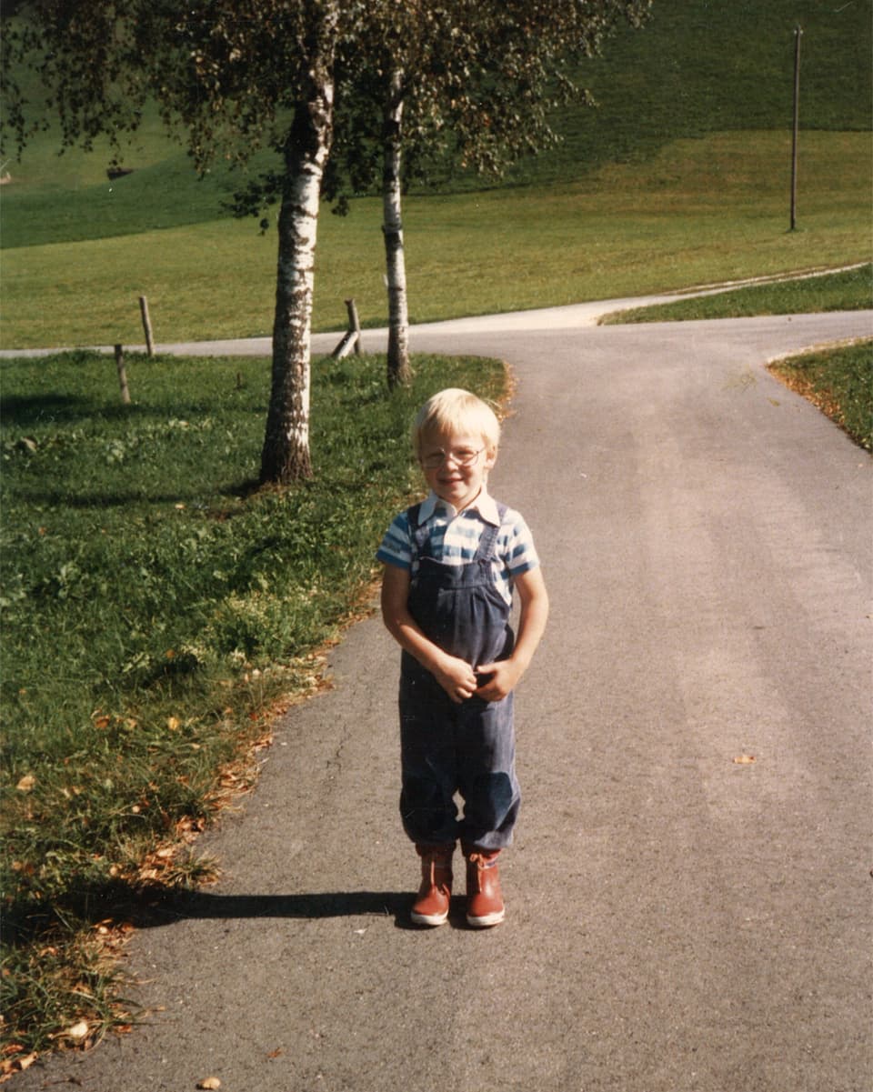 Ein Bub mit gestreiftem T-Shirt, blauer Latzhose und roten Gummistiefeln steht mitten auf einer Landstrasse.