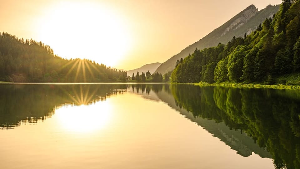 Sonnenaufgang über einem ruhigen See, umgeben von Bergen und Wäldern.