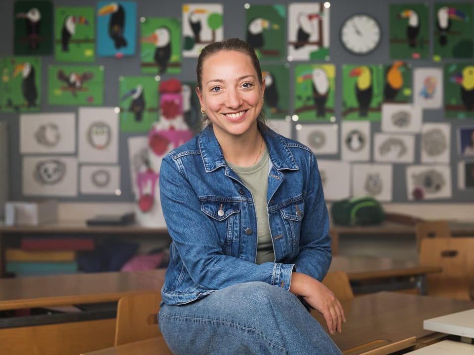 Moderatorin Anna Zöllig sitzt auf einer Schulbank in einem Klassenzimmer.