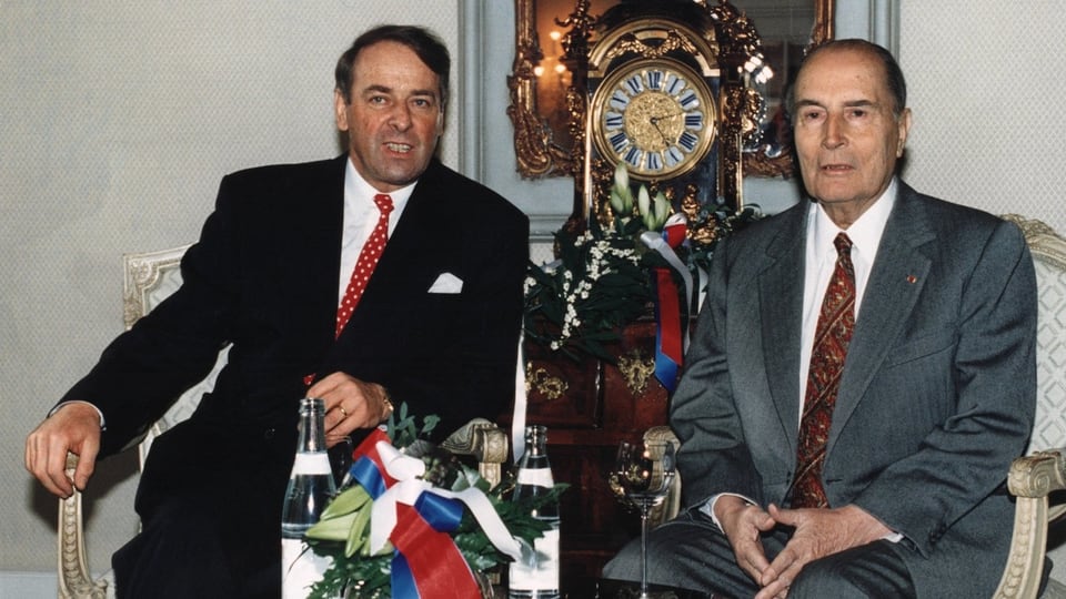 Adolf Ogi und François Mitterrand sitzen nebeneinander bei einem Glas Wasser.
