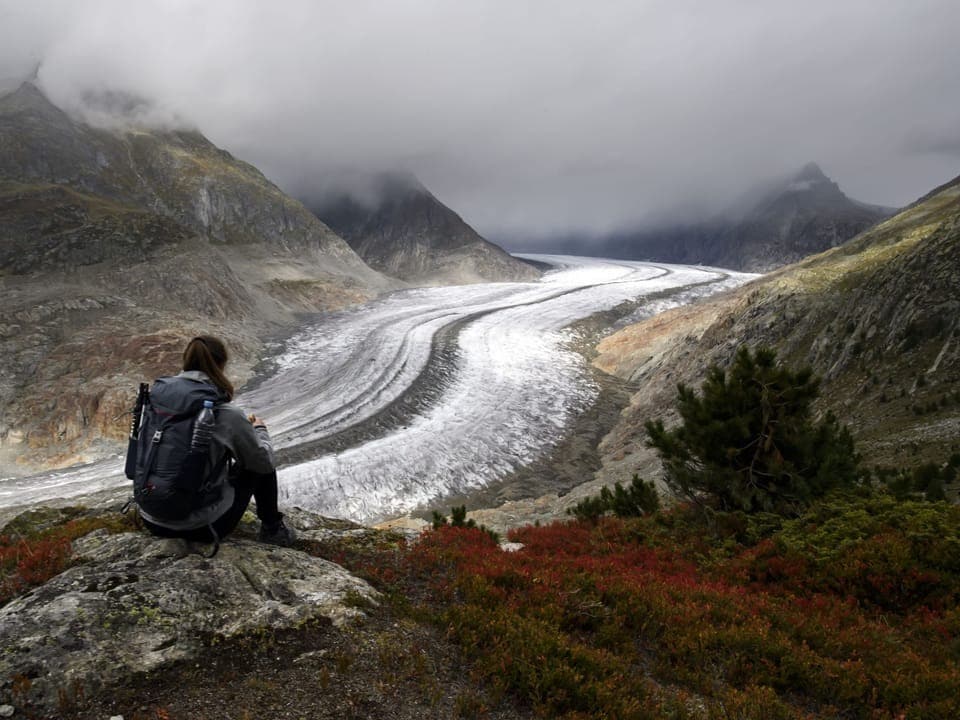 Eine Wanderin sitzt an einem nebligen Herbsttag auf einem Stein mit Blick auf den schmelzenden Aletschgletscher.