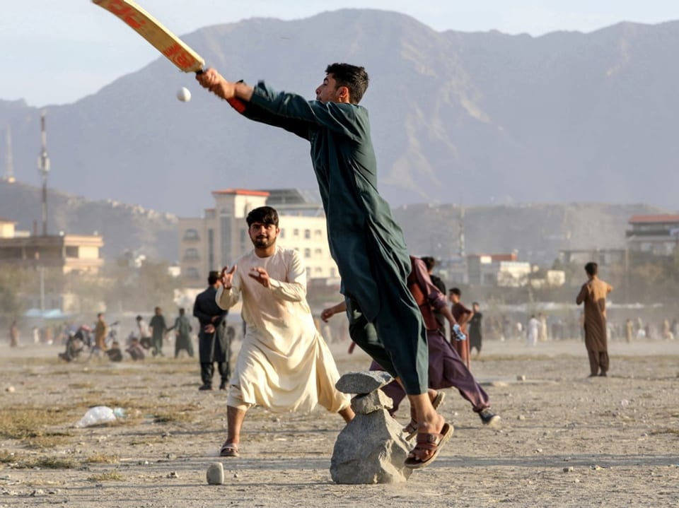 Männer spielen auf staubigem Feld Cricket vor Bergkulisse.