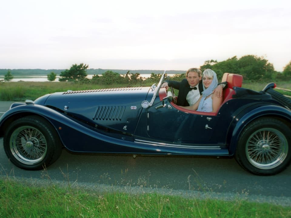 Paar in einem klassischen, offenen Oldtimer auf einer Strasse.