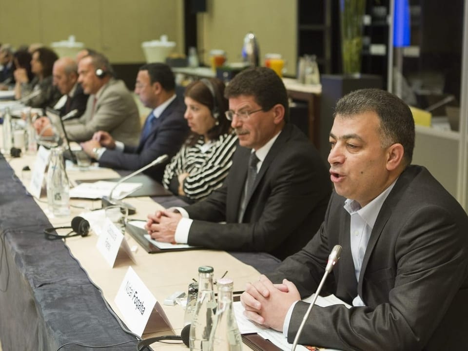 Mann spricht während einer Konferenz, mehrere Teilnehmer sitzen am Tisch.
