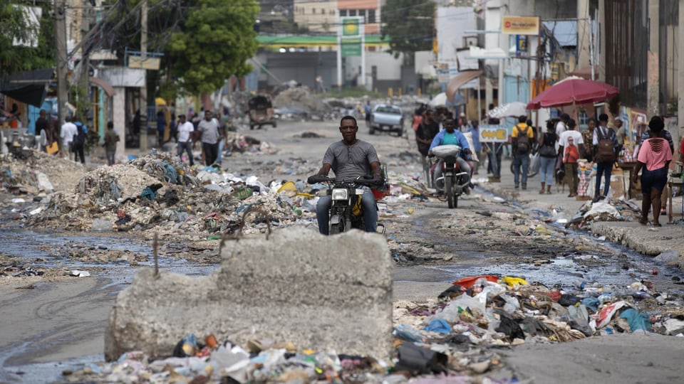 Menschen und Motorräder in einer mit Müll übersäten Strasse in Port-Au-Prince.