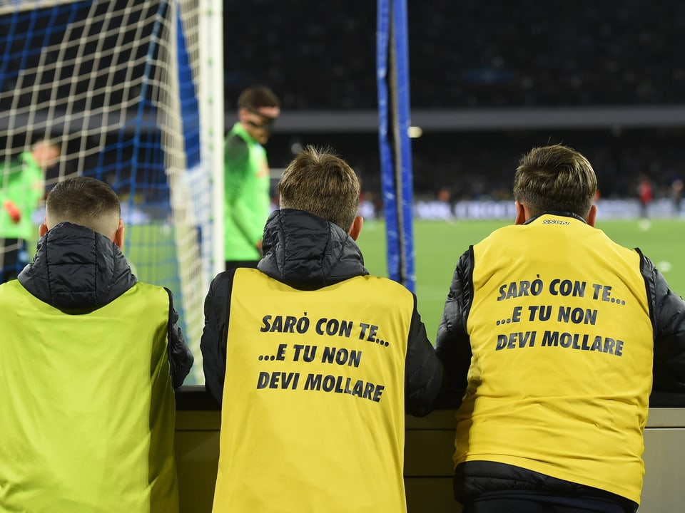 Drei Menschen in gelben Westen mit Aufschrift stehen vor einem Fussballtor.